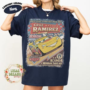 May include: Navy blue t-shirt featuring a vintage-style graphic of Cruz Ramirez from the Disney Pixar Cars movie. The design includes the text "CRUZ RAMIREZ" and "ROUND 4 WINNER TAKES ALL". The shirt is from Lucas Dreams Boutique.