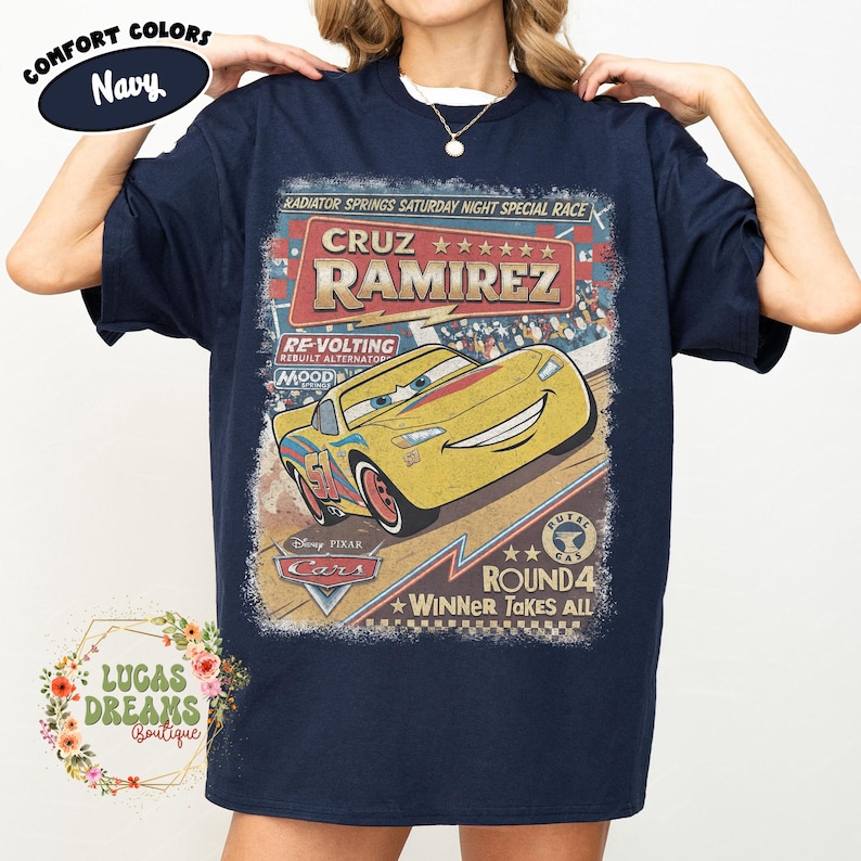 May include: Navy blue t-shirt featuring a vintage-style graphic of Cruz Ramirez from the Disney Pixar Cars movie. The design includes the text "CRUZ RAMIREZ" and "ROUND 4 WINNER TAKES ALL". The shirt is from Lucas Dreams Boutique.