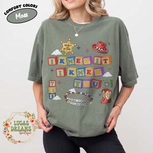 May include: A moss green short-sleeve t-shirt featuring a graphic print of colorful alphabet blocks spelling I KNEW IT I KNEW YOU. The design includes a sheriff star, a red cowboy hat, a pair of cowboy boots, and a beaded bracelet with text.