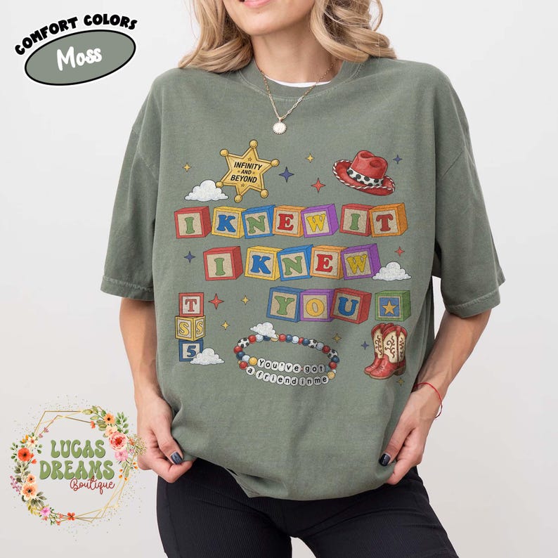 May include: A moss green short-sleeve t-shirt featuring a graphic print of colorful alphabet blocks spelling I KNEW IT I KNEW YOU. The design includes a sheriff star, a red cowboy hat, a pair of cowboy boots, and a beaded bracelet with text.