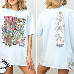May include: Light blue t-shirt featuring the words "Toy Story" in pink and yellow, with various cartoon characters. The back of the shirt has a vertical design of characters and stars. The shirt is a comfortable, casual style.
