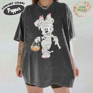 May include: Dark gray t-shirt featuring a Halloween-themed Minnie Mouse design. Minnie is depicted as a mummy holding a jack-o'-lantern basket. The shirt has the text "Comfort Colors Pepper" on the upper left.