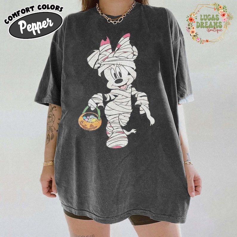 May include: Dark gray t-shirt featuring a Halloween-themed Minnie Mouse design. Minnie is depicted as a mummy holding a jack-o'-lantern basket. The shirt has the text "Comfort Colors Pepper" on the upper left.