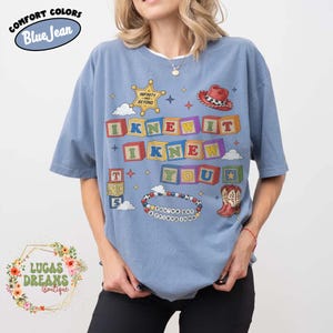 May include: A blue short-sleeve t-shirt featuring a graphic print of colorful alphabet blocks spelling I KNEW IT I KNEW YOU, a sheriff badge with the text INFINITY AND BEYOND, a red cowboy hat, a pair of cowboy boots, and a beaded bracelet with text.