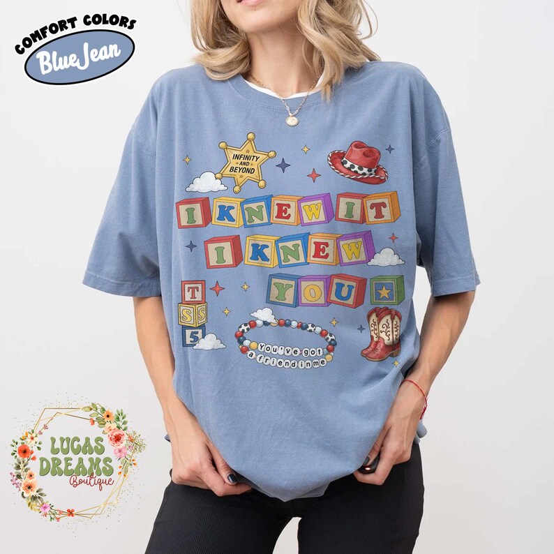 May include: A blue short-sleeve t-shirt featuring a graphic print of colorful alphabet blocks spelling I KNEW IT I KNEW YOU, a sheriff badge with the text INFINITY AND BEYOND, a red cowboy hat, a pair of cowboy boots, and a beaded bracelet with text.