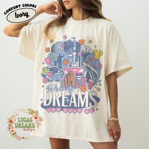 K&ouml;nnte beinhalten: Elfenbeinfarbenes T-Shirt mit einem farbenfrohen Grafikdesign mit verschiedenen Themenpark-Symbolen und dem Text "What Dreams Are Made Of". Das Design umfasst Blumen, Sterne und Herzformen. Das Shirt ist von der Marke Comfort Colors.