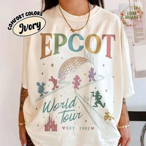 Puede incluir: Camiseta de color marfil con un dise&ntilde;o gr&aacute;fico colorido que presenta la palabra "EPCOT" y "World Tour" con el a&ntilde;o "EST. 1982". El dise&ntilde;o incluye personajes de dibujos animados y una c&uacute;pula geom&eacute;trica. La camiseta tiene un corte holgado.