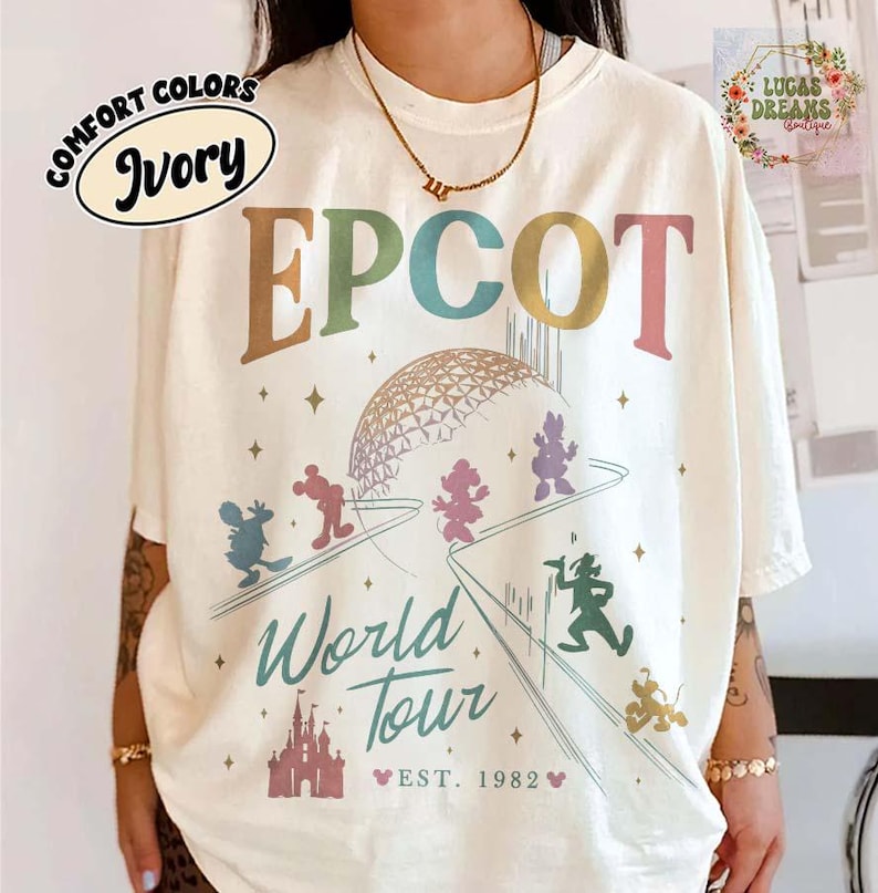 Puede incluir: Camiseta de color marfil con un dise&ntilde;o gr&aacute;fico colorido que presenta la palabra "EPCOT" y "World Tour" con el a&ntilde;o "EST. 1982". El dise&ntilde;o incluye personajes de dibujos animados y una c&uacute;pula geom&eacute;trica. La camiseta tiene un corte holgado.