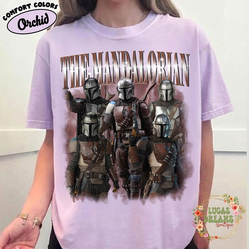 Puede incluir: Una camiseta de manga corta de color morado claro con un estampado gr&aacute;fico de cinco personajes Mandalorianos con armadura met&aacute;lica. El texto THE MANDALORIAN est&aacute; impreso en una fuente grande, negrita y de tonos marrones sobre los personajes.