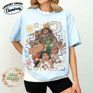 May include: A light blue t-shirt featuring a colorful graphic print of various animated characters, including a central figure with a large hook, a rooster, and a pig. The design includes decorative sun, wave, and spiral patterns in earthy tones.