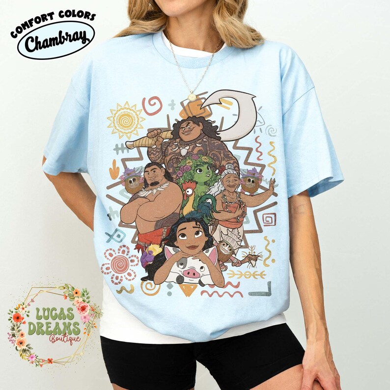 May include: A light blue t-shirt featuring a colorful graphic print of various animated characters, including a central figure with a large hook, a rooster, and a pig. The design includes decorative sun, wave, and spiral patterns in earthy tones.