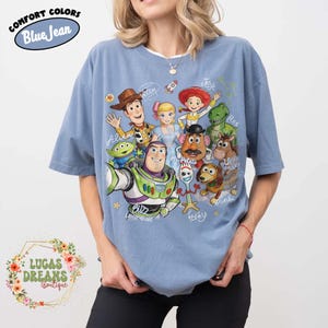 Op de afbeelding: Een jeansblauw t-shirt met korte mouwen en een grafische print van Toy Story-personages, waaronder Woody, Buzz Lightyear, Jessie, Rex, Slinky Dog, Mr. Potato Head, Bo Peep, Forky en een alien, met handgeschreven namen van de personages.