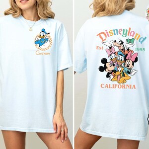 Puede incluir: Camiseta azul claro con Donald Duck en la parte delantera y un dise&ntilde;o de Disneylandia en la parte trasera. El dise&ntilde;o de Disneylandia incluye a Mickey Mouse, Minnie Mouse, Goofy, Donald Duck y Pluto. La camiseta tiene el texto "Disneyland Est 1955 California".