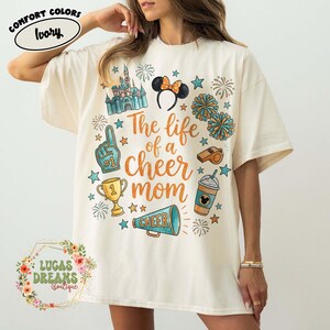 Puede incluir: Camiseta de color marfil con un diseño gráfico colorido. El diseño incluye un castillo, orejas de Minnie Mouse, pompones, un trofeo y el texto "The life of a cheer mom". La camiseta tiene mangas cortas y un corte holgado.