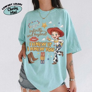 May include: A light mint green t-shirt featuring a graphic of Jessie from Toy Story, a red cowboy hat, brown boots, and alphabet blocks. The text reads "infinity and beyond" and "I KNEW IT I KNEW YOU" in a stylized brown font with small stars.