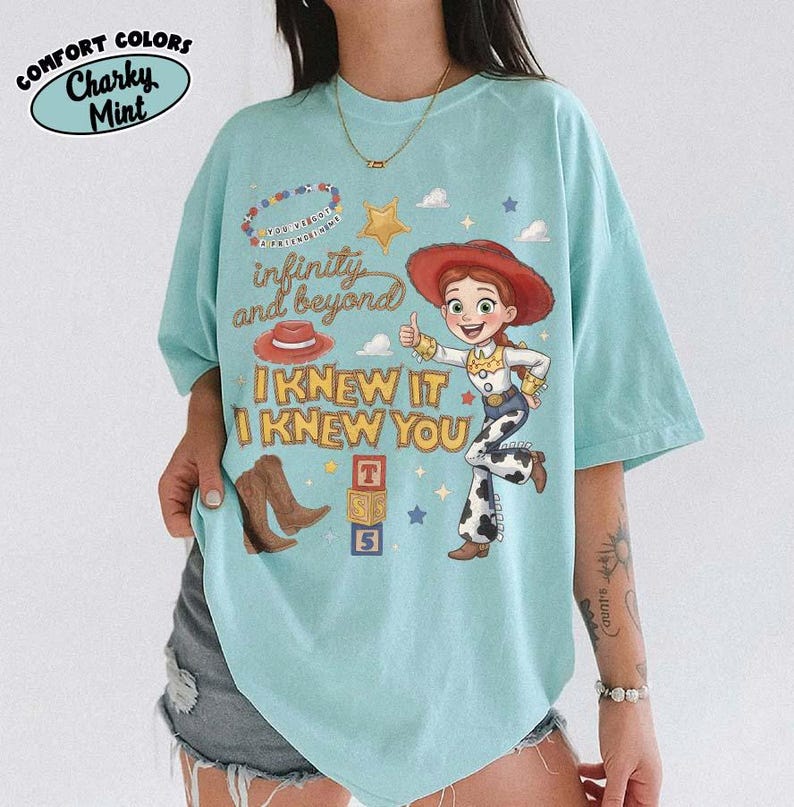 May include: A light mint green t-shirt featuring a graphic of Jessie from Toy Story, a red cowboy hat, brown boots, and alphabet blocks. The text reads "infinity and beyond" and "I KNEW IT I KNEW YOU" in a stylized brown font with small stars.
