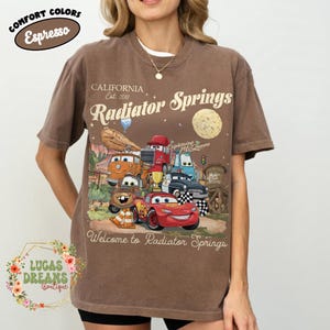 Pode incluir: Camiseta marrom com um gr&aacute;fico de personagens do filme Carros, incluindo Rel&acirc;mpago McQueen. A camisa tem o texto "Radiator Springs" e "Welcome to Radiator Springs". A camisa tamb&eacute;m tem o texto "Comfort Colors Espresso".