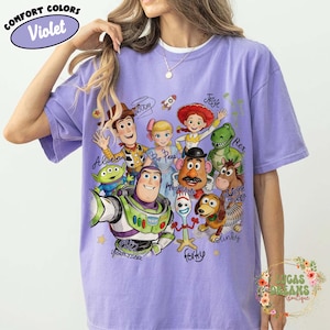 Op de afbeelding: Een violetkleurig T-shirt met korte mouwen en een grafische print van Toy Story-personages, waaronder Buzz Lightyear, Woody, Bo Peep, Jessie, Rex, Mr. Potato Head, Bullseye, Slinky Dog, Forky en een Alien, met hun namen in cursief schrift.