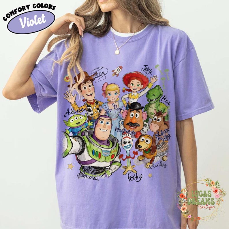 Op de afbeelding: Een violetkleurig T-shirt met korte mouwen en een grafische print van Toy Story-personages, waaronder Buzz Lightyear, Woody, Bo Peep, Jessie, Rex, Mr. Potato Head, Bullseye, Slinky Dog, Forky en een Alien, met hun namen in cursief schrift.