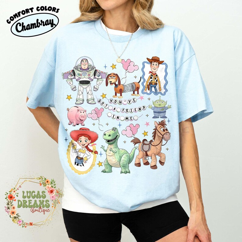 May include: Light blue t-shirt featuring cartoon characters including Buzz Lightyear, Woody, and Rex. The shirt has the phrase "YOU'VE GOT A FRIEND IN ME" and the Comfort Colors and Chambray logos. The shirt is from Lucas Dreams Boutique.