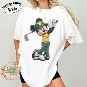 May include: White t-shirt featuring a cartoon Mickey Mouse in a golf outfit, swinging a golf club. The design includes a green hat, yellow shirt, and green pants. The text "COMFORT COLORS White" is at the top, and "LUCAS DREAMS Boutique" is at the bottom.