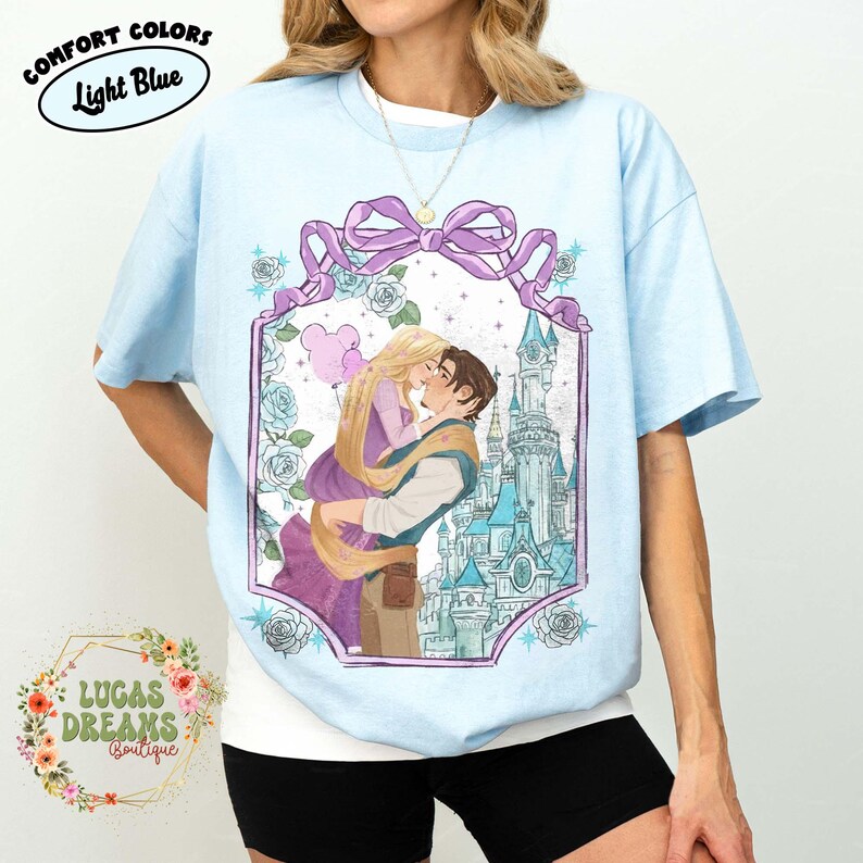 Puede incluir: Camiseta azul claro con un gr&aacute;fico de una pareja bes&aacute;ndose frente a un castillo. El gr&aacute;fico est&aacute; enmarcado por un lazo morado y rosas. El texto en la camiseta dice "Comfort Colors" y "Light Blue". El logotipo de la boutique tambi&eacute;n es visible.