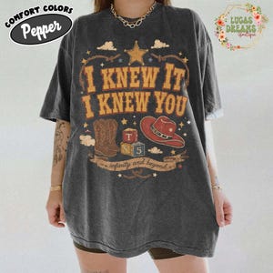 May include: A charcoal grey short-sleeve t-shirt featuring a graphic print with the text I KNEW IT I KNEW YOU, a star, a cowboy boot, a red cowboy hat, and two blocks labelled T and S 5, with the text infinity and beyond on a banner below.