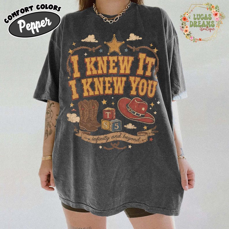 May include: A charcoal grey short-sleeve t-shirt featuring a graphic print with the text I KNEW IT I KNEW YOU, a star, a cowboy boot, a red cowboy hat, and two blocks labelled T and S 5, with the text infinity and beyond on a banner below.