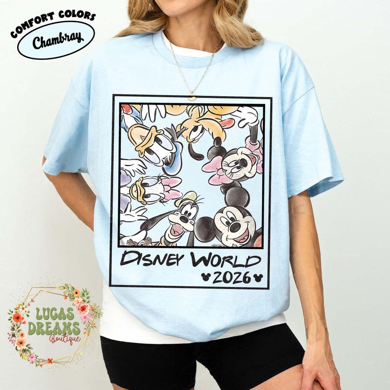 Op de afbeelding: Lichtblauw T-shirt met een omlijnde afbeelding van Disney-personages, waaronder Mickey en Minnie Mouse, Donald Duck en Goofy. De tekst "DISNEY WORLD 2026" staat eronder. Het shirt heeft de Comfort Colors en Chambray logo's.