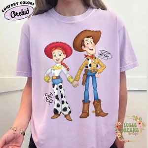 Puede incluir: Una camiseta de color lila claro con una ilustraci&oacute;n de dibujos animados de Jessie y Woody de Toy Story. Jessie lleva un sombrero rojo, una camisa blanca y pantalones de lunares blancos y negros. Woody lleva un sombrero marr&oacute;n, una camisa amarilla y vaqueros azules. La camiseta tiene el texto "Comfort Colors Orchid".