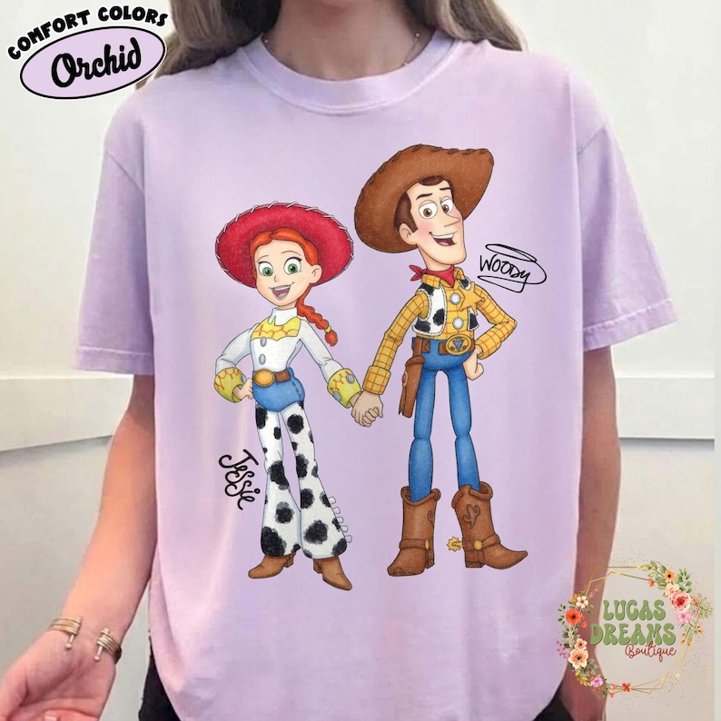 Puede incluir: Una camiseta de color lila claro con una ilustraci&oacute;n de dibujos animados de Jessie y Woody de Toy Story. Jessie lleva un sombrero rojo, una camisa blanca y pantalones de lunares blancos y negros. Woody lleva un sombrero marr&oacute;n, una camisa amarilla y vaqueros azules. La camiseta tiene el texto "Comfort Colors Orchid".