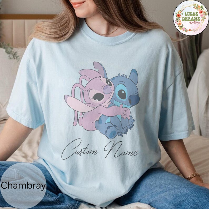 May include: Light blue t-shirt featuring a cartoon illustration of two characters hugging. One character is blue, the other is pink. The words "Custom Name" are written in cursive below the illustration. The word "Chambray" is at the bottom left.