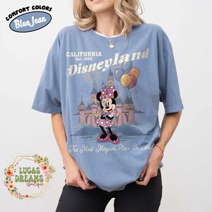 May include: Blue t-shirt with a Disneyland graphic. The design features Minnie Mouse in a pink dress and bow, in front of a castle, with balloons and fireworks. Text includes "CALIFORNIA Disneyland Est. 1955" and "The Most Magical Place On Earth."