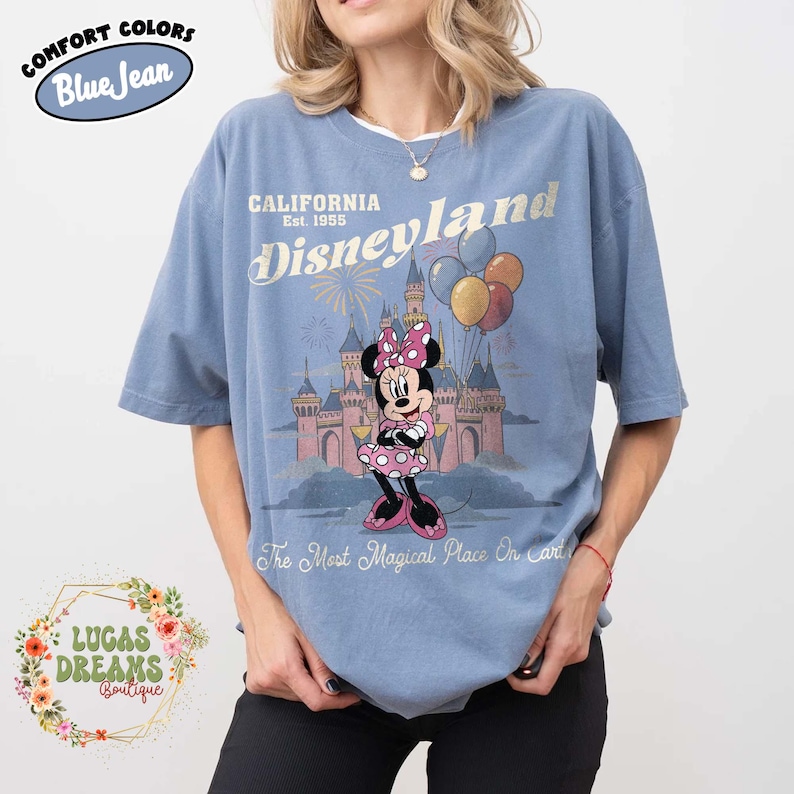May include: Blue t-shirt with a Disneyland graphic. The design features Minnie Mouse in a pink dress and bow, in front of a castle, with balloons and fireworks. Text includes "CALIFORNIA Disneyland Est. 1955" and "The Most Magical Place On Earth."