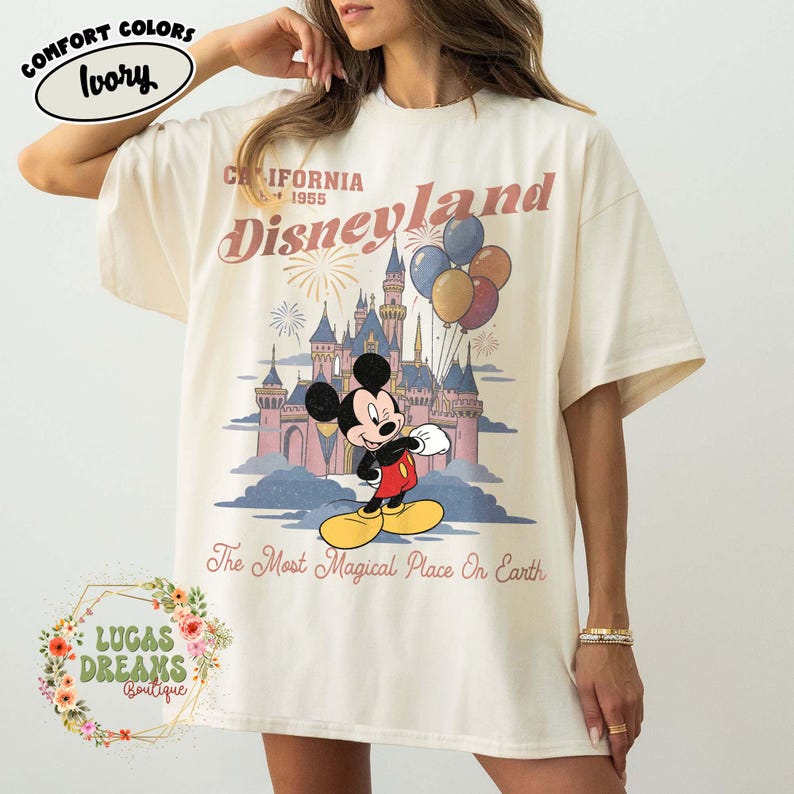 May include: Ivory-colored t-shirt featuring a Disneyland graphic with Mickey Mouse, a castle, and balloons. The text reads "California 1955 Disneyland" and "The Most Magical Place On Earth". The shirt is from Lucas Dreams Boutique.