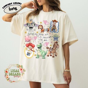 May include: Ivory-colored Comfort Colors t-shirt featuring colorful cartoon characters and the phrase "YOU'VE GOT A FRIEND IN ME." The design includes Buzz Lightyear, Woody, and other Toy Story characters.