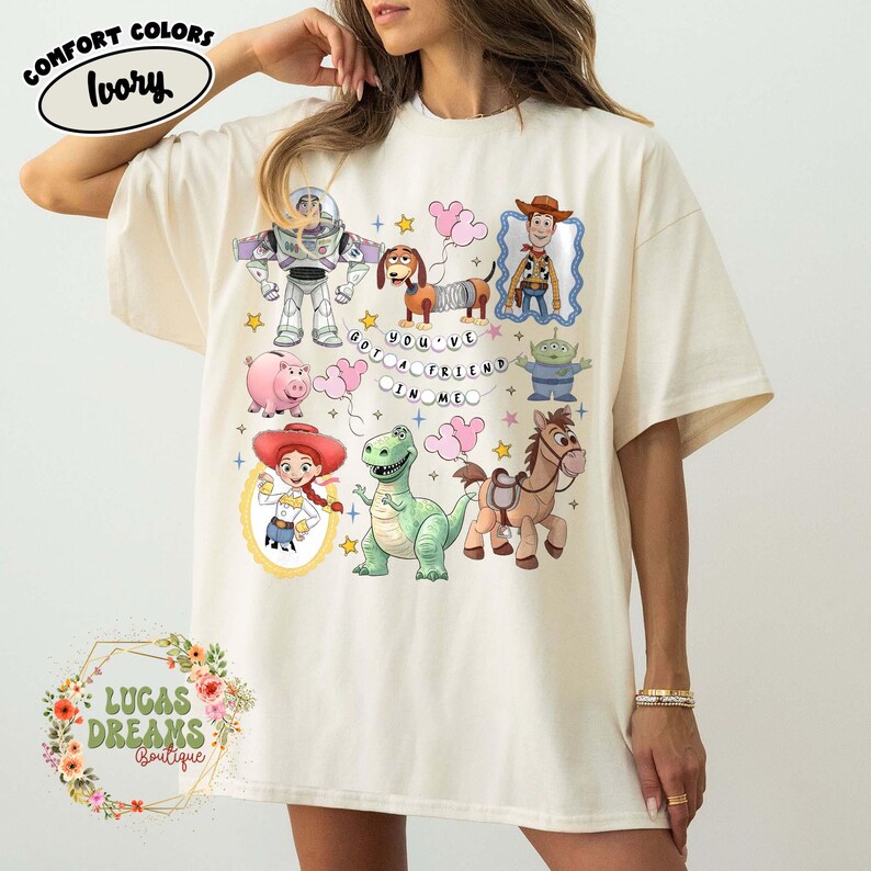 May include: Ivory-colored Comfort Colors t-shirt featuring colorful cartoon characters and the phrase "YOU'VE GOT A FRIEND IN ME." The design includes Buzz Lightyear, Woody, and other Toy Story characters.