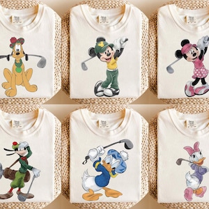 May include: Six off-white t-shirts featuring Disney characters playing golf. Pluto, Mickey Mouse, Minnie Mouse, Goofy, Donald Duck, and Daisy Duck are depicted in colorful cartoon graphics, each holding a golf club.