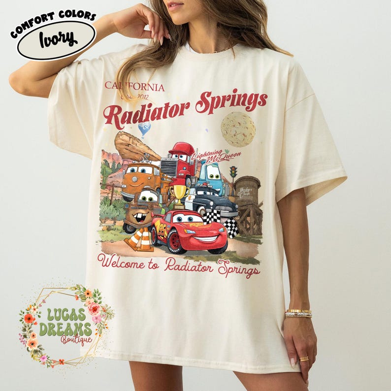 Pode incluir: Camiseta cor marfim com um gr&aacute;fico de personagens do filme Carros, incluindo o Rel&acirc;mpago McQueen. O design inclui o texto "Radiator Springs" e "Welcome to Radiator Springs". A camiseta tem mangas curtas e um corte folgado.