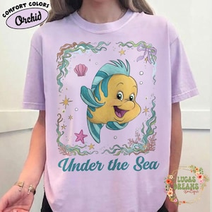 May include: A light purple t-shirt featuring a cartoon image of Flounder from The Little Mermaid, surrounded by aquatic elements. The text "Under the Sea" is printed below the image. The shirt also has the text "Comfort Colors Orchid" printed on it.