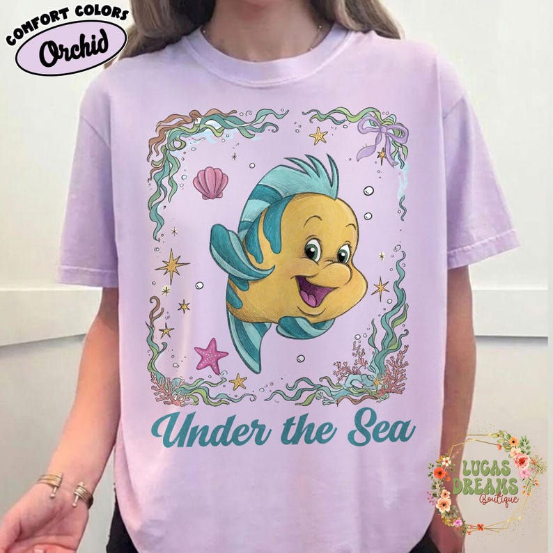 May include: A light purple t-shirt featuring a cartoon image of Flounder from The Little Mermaid, surrounded by aquatic elements. The text "Under the Sea" is printed below the image. The shirt also has the text "Comfort Colors Orchid" printed on it.