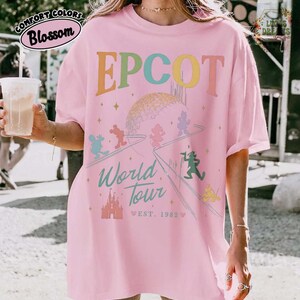 Puede incluir: Camiseta rosa de EPCOT World Tour con un gr&aacute;fico colorido con Mickey Mouse y otros personajes de Disney. La camiseta tiene el texto "EPCOT" en colores arco&iacute;ris, un gr&aacute;fico de Spaceship Earth y "World Tour" con "Est. 1982".