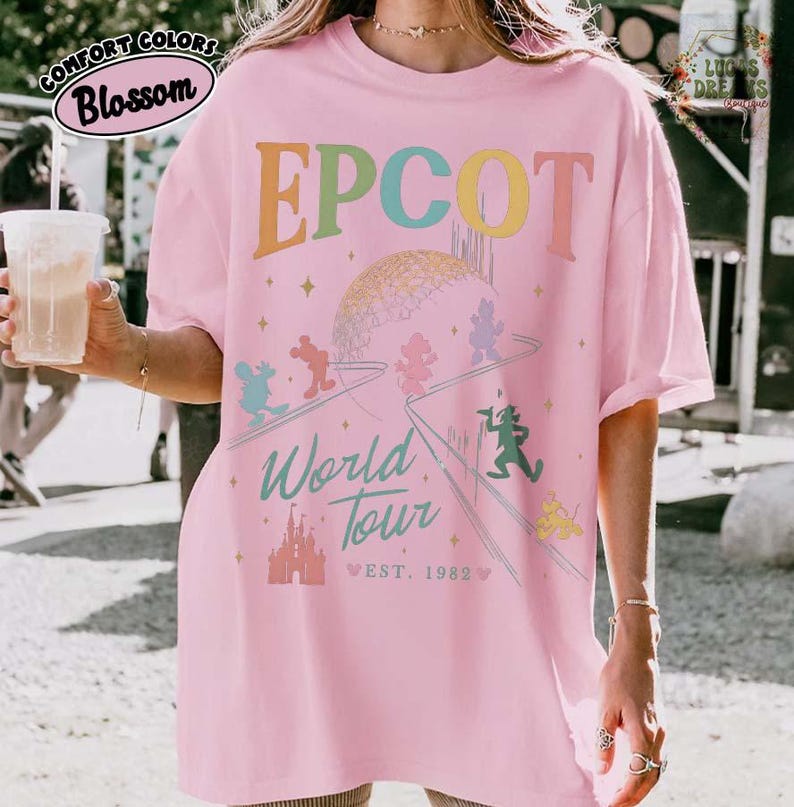 Puede incluir: Camiseta rosa de EPCOT World Tour con un gr&aacute;fico colorido con Mickey Mouse y otros personajes de Disney. La camiseta tiene el texto "EPCOT" en colores arco&iacute;ris, un gr&aacute;fico de Spaceship Earth y "World Tour" con "Est. 1982".
