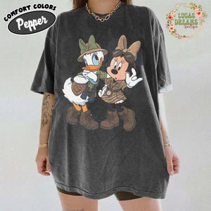 May include: Dark grey t-shirt with a graphic of Donald Duck and Minnie Mouse in safari outfits. The shirt has the words "COMFORT COLORS" and "Pepper" in a circular design on the upper left. The words "LUCAS DREAMS boutique" are in a floral design on the upper right.