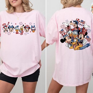 以下が含まれることがあります： 愛国的な漫画のデザインが施された、明るいピンク色のTシャツ。前面には、お祝いの帽子と花火をかぶった漫画のキャラクターが描かれています。Tシャツの背面には、アメリカの国旗と花火を持ったキャラクターのグループが描かれています。