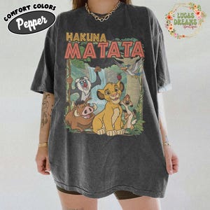 May include: Dark gray t-shirt featuring the phrase "Hakuna Matata" above a colorful graphic of characters from The Lion King. The shirt has a vintage, slightly faded look. The shirt is a Comfort Colors Pepper brand.