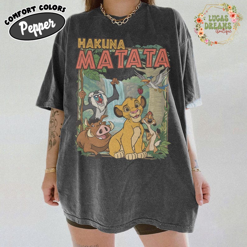 May include: Dark gray t-shirt featuring the phrase "Hakuna Matata" above a colorful graphic of characters from The Lion King. The shirt has a vintage, slightly faded look. The shirt is a Comfort Colors Pepper brand.