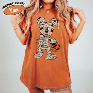May include: An orange t-shirt featuring a cartoon mummy character holding a jack-o'-lantern bucket. The character is wrapped in bandages and has a smiling face. The shirt has the words "Comfort Colors" and "Yam" printed on it.