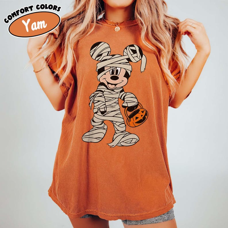 May include: An orange t-shirt featuring a cartoon mummy character holding a jack-o'-lantern bucket. The character is wrapped in bandages and has a smiling face. The shirt has the words "Comfort Colors" and "Yam" printed on it.