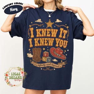 May include: A navy blue t-shirt featuring a graphic print with the text I KNEW IT I KNEW YOU and infinity and beyond. The design includes a gold star, a pair of brown cowboy boots, a red cowboy hat, and two alphabet blocks labelled T and S 5.